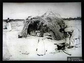 Mujer y niña ¿indígenas? al lado de una hoguera y una cabaña